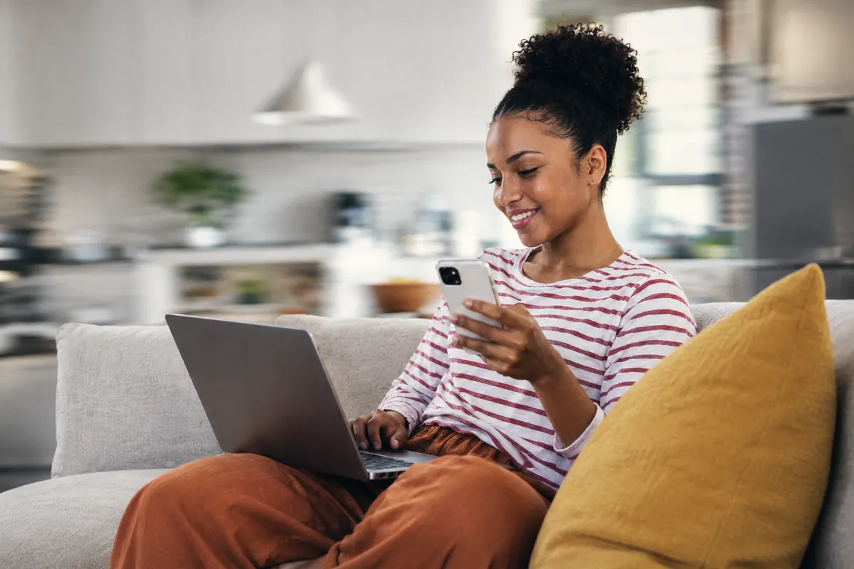 A woman on her sofa looking at her phone with a laptop open